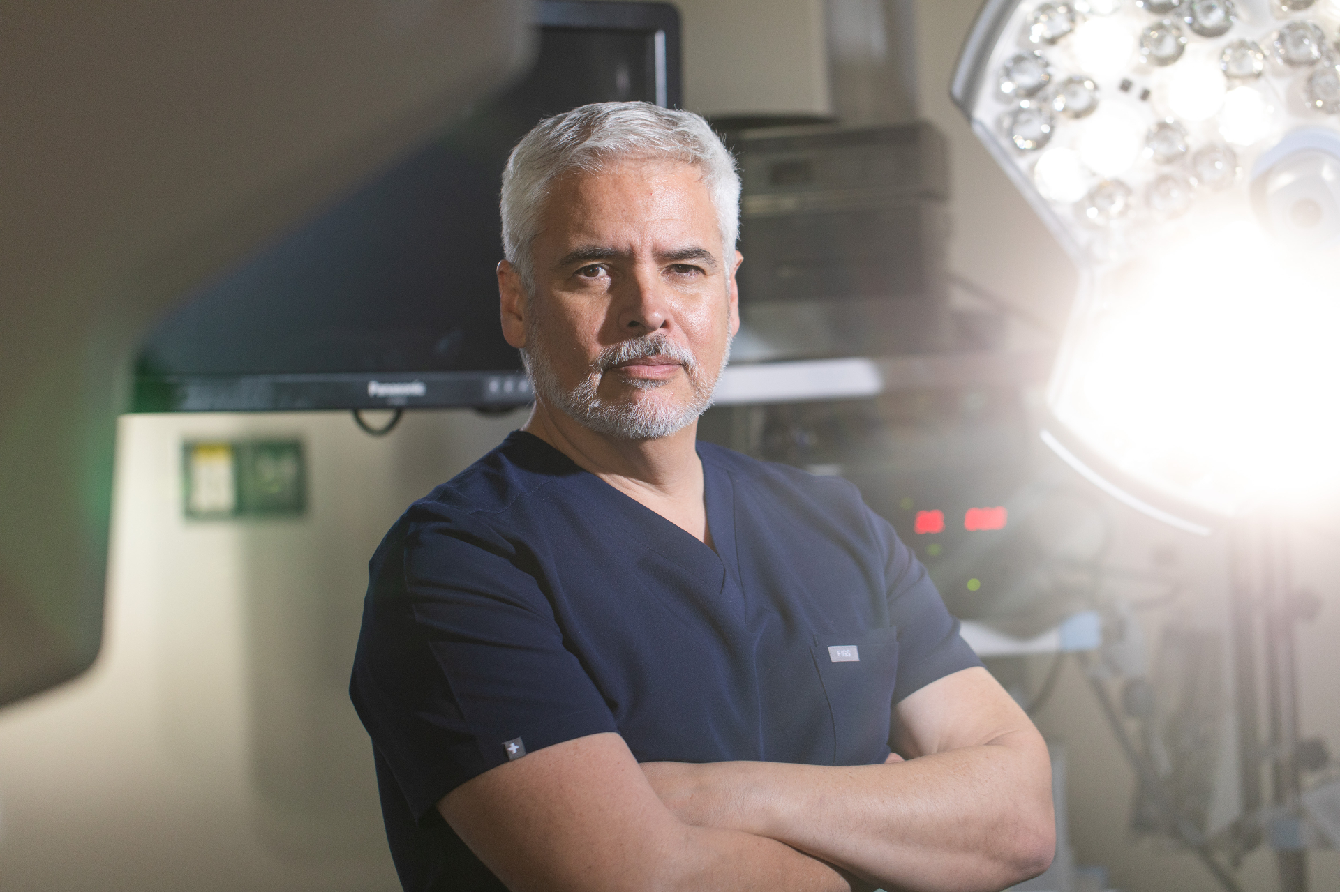 Portrait of Dr. Ariel Ortiz in scrubs standing in an operating room beside a surgical light.